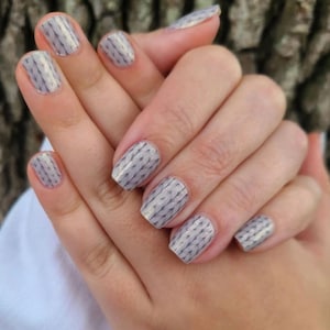 May include: Close-up of a hand with light gray nail polish. Each nail is decorated with a knitted pattern in a darker gray color. The nails are short and square-shaped.