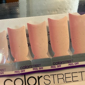 May include: A package of Color Street nail polish strips. The strips are a light pink color with a speckled texture. The package has the words "Color Street" and "Peel Off" printed on it.