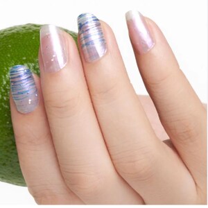 May include: Close-up of a hand with manicured nails. The nails feature a pearlescent finish with a subtle pink hue. Some nails are adorned with blue and silver striped designs, while others have a clear, shimmering appearance. A green lime is visible in the background.
