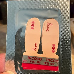 May include: A package containing nail polish strips. The strips are light pink with the word "love" written on them in red script. Small red hearts are also on the strips. The package has the brand name "INCOCO" printed on it.