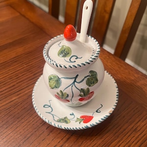 May include: A white ceramic sugar bowl with a matching saucer. The bowl and saucer are decorated with a hand-painted strawberry and leaf design. A white spoon rests in the bowl, and the lid is topped with a red strawberry.