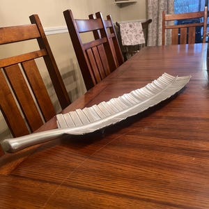 May include: A silver-colored, leaf-shaped decorative tray. The tray is made of metal and has a detailed, textured surface. It is placed on a dark wooden table, with wooden chairs in the background. The tray is approximately 24 inches long.