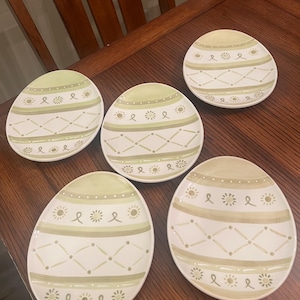 May include: Five egg-shaped decorative plates with a white base and green and tan painted designs. Each plate features a different pattern of stripes, dots, and floral accents. The plates are arranged on a dark wooden surface.