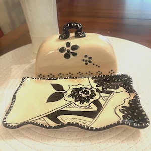 May include: A beige ceramic butter dish with a black handle and floral accents. The matching rectangular serving dish features a black and white floral design. The set is placed on a white, textured surface.