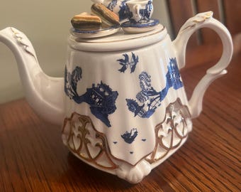 May include: A white teapot with a blue and white willow pattern and gold trim. The teapot has a lid with a miniature teapot, cup, and saucer on top. The teapot is sitting on a wooden table.