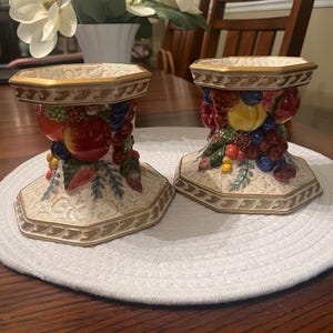 May include: Two octagonal ceramic candle holders featuring a raised, colorful fruit motif including apples, grapes, and berries. The base and top rims are accented with gold-toned trim, and the cream-colored body displays a subtle embossed pattern.