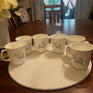 May include: A set of six off-white ceramic mugs, each featuring a delicate floral pattern with small orange and yellow flowers and green leaves. The mugs have a smooth, rounded shape with a single handle on the side, arranged in a circular formation.