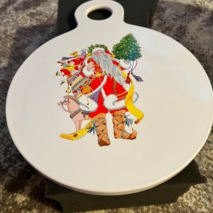 May include: White ceramic round serving tray with a handle. The tray features a colorful illustration of Santa Claus carrying a sack of gifts, a Christmas tree, and a rocking horse.