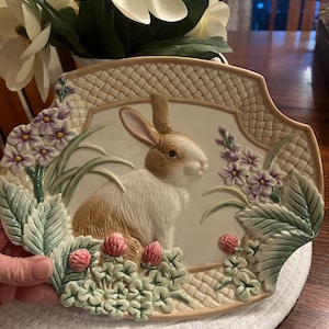 May include: A decorative ceramic plate featuring a detailed relief of a brown and white rabbit surrounded by flowers and foliage. The plate has a woven border and is in a rectangular shape with a scalloped edge. The color palette includes cream, brown, green, and purple.