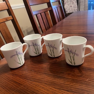 May include: Four white ceramic mugs with a curved handle, each adorned with a purple iris flower design. The mugs are arranged on a dark wooden table, suggesting a dining or kitchen setting. The mugs are approximately 3 inches tall.