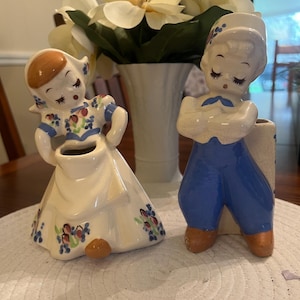 May include: Two ceramic figurines of a Dutch boy and girl. The girl wears a white dress with blue and floral accents. The boy wears blue pants and a white shirt. Both have closed eyes and are holding vases.