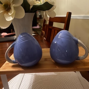 May include: A pair of periwinkle blue ceramic salt and pepper shakers, each featuring a rounded, teardrop-like body with a curved handle on the side and small holes at the top for dispensing seasoning, presented as a matching set of two.