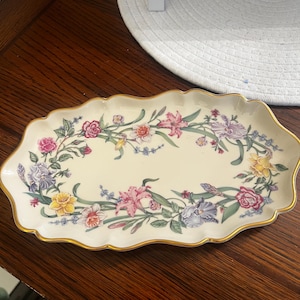 May include: An oval ceramic tray with a scalloped edge and a gold-trimmed rim. The center features a cream-colored background decorated with a continuous floral garland pattern consisting of pink, yellow, purple, and white flowers with green leaves.