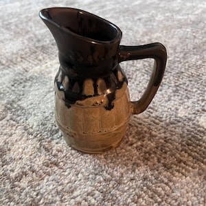 May include: A small, brown and tan ceramic pitcher with a wooden barrel design. The pitcher has a black glaze and a handle.