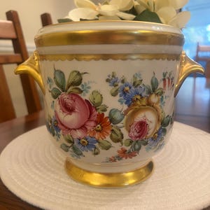 May include: A white and gold ceramic vase with floral designs. The vase features pink, blue, and orange flowers with green leaves. Gold trim accents the top, base, and handles. The vase is sitting on a white circular placemat.