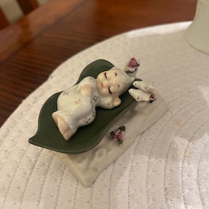 May include: A porcelain figurine of a sleeping baby, lying on a green leaf-shaped base atop a pillow. The baby is wearing a white outfit with gold accents. Small pink roses are placed on the pillow.