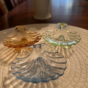May include: Three decorative glass seashell-shaped dishes in shades of orange, green, and blue. Each dish has a scalloped edge and sits on a small pedestal base. The dishes are arranged on a textured, off-white surface.