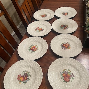 May include: Eight white ceramic plates with a textured, dotted rim and a central fruit design. The fruit design includes peaches, grapes, and other fruits in shades of orange, red, and purple. The plates are arranged on a dark wooden table.