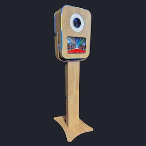 May include: A wooden photo booth with a rectangular head and a tall, rectangular base. The head has a circular light and a screen displaying a red carpet scene. The booth is illuminated with blue and yellow lights.