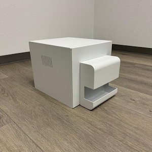 May include: A white, modern-style box with a rectangular main body and a smaller, curved extension. The main body has a series of vertical slits on one side. The object is set on a wooden floor.