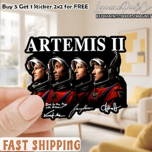 May include: A sticker featuring the text "ARTEMIS II" above four astronaut portraits in red and black space suits. The sticker also includes the text "Back to the Moon with Artemis" and signatures. The sticker is held by a person's hand. The top of the sticker reads "Buy 5 Get 1 Sticker 2x2 for FREE".
