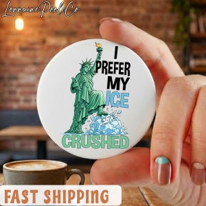 May include: A white round button with the text "I PREFER MY ICE CRUSHED" and an illustration of the Statue of Liberty. The Statue of Liberty is green and is standing on a pile of crushed ice. The button is held by a hand.