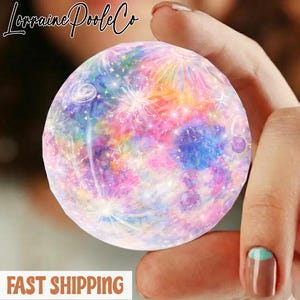 May include: A round, colorful decorative object with a galaxy design. The object features a blend of pink, blue, yellow, and purple hues, with white starburst patterns. The text "Lorraine Poole Co" is at the top.