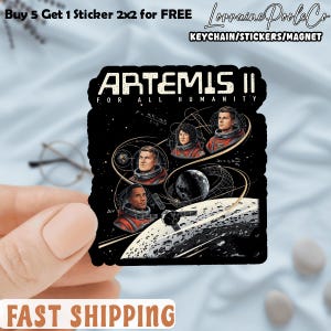 May include: Black sticker featuring the text "ARTEMIS II FOR ALL HUMANITY" and images of four astronauts in red suits. The sticker also includes a depiction of a planet, spacecraft, and scientific diagrams. The sticker is held by a hand.