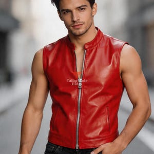 May include: A sleeveless, red leather vest with a silver zipper closure. The vest has a stand-up collar and two front pockets. The model is wearing black jeans. The vest is from TailorMade Hide.