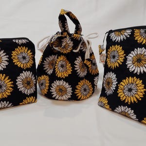 May include: Three black fabric bags with a repeating pattern of yellow and white daisies. The bags include a small zippered pouch, a drawstring bag, and a larger zippered pouch. The bags are likely for cosmetics or small items.