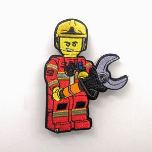 May include: Embroidered patch of a firefighter in a red uniform and yellow helmet, holding a silver and black tool. The patch has a cartoon design with a smiling face.