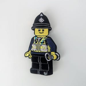 May include: An embroidered patch of a Lego police officer. The officer is wearing a blue uniform, a yellow vest, a black belt, and a black helmet with a silver badge. The officer is holding a pair of handcuffs in one hand.