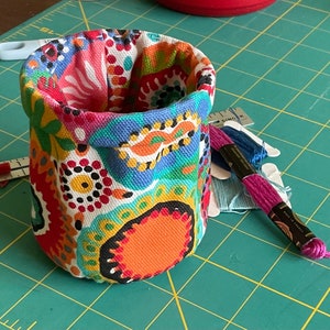 May include: A cylindrical fabric container with a colorful floral pattern, featuring red, orange, blue, and yellow hues. Embroidery floss in shades of blue and pink rests beside the container, along with a small ruler. The container is likely for craft supplies.