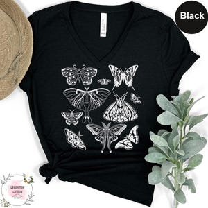 a black t-shirt with a white graphic design of various butterflies printed on it. The t-shirt is placed on a white surface, and there is a hat and some greenery visible in the background.