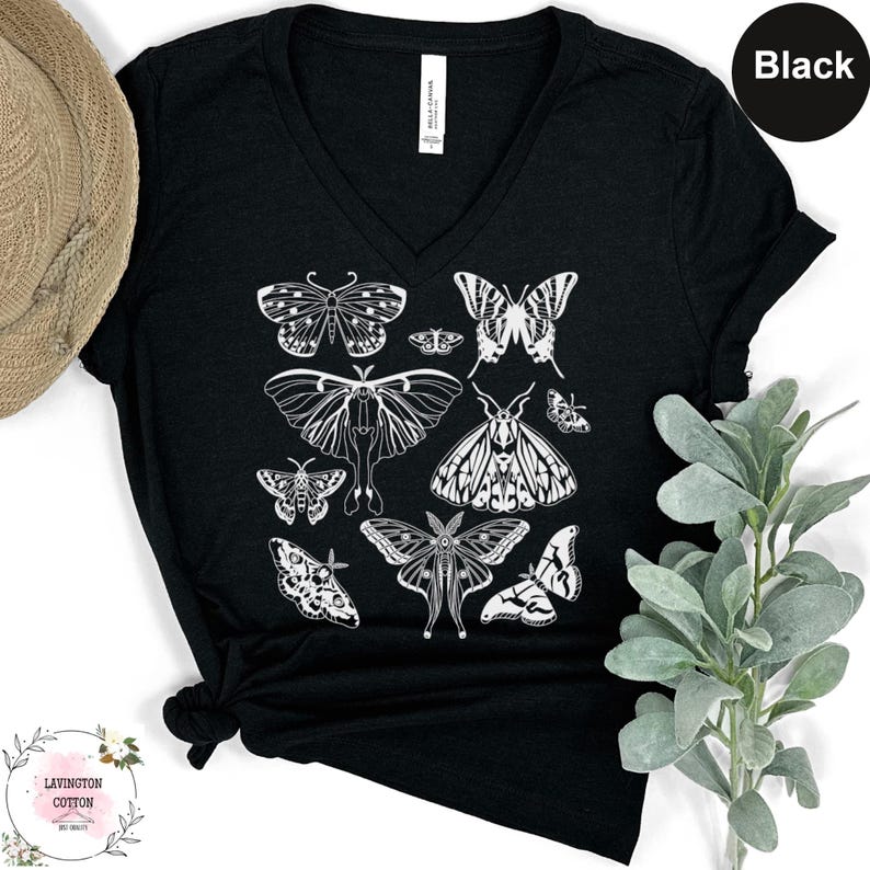 a black t-shirt with a white graphic design of various butterflies printed on it. The t-shirt is placed on a white surface, and there is a hat and some greenery visible in the background.
