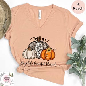 May include: Peach-colored V-neck t-shirt featuring a pumpkin graphic. The graphic includes three pumpkins, one with a leopard print, and the words "grateful thankful blessed" in a script font. The shirt is made of soft material.
