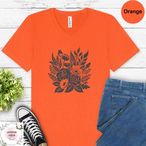 a bright orange t-shirt with a black floral design, placed on a white wooden surface alongside a pair of blue jeans and a pair of black and white sneakers.