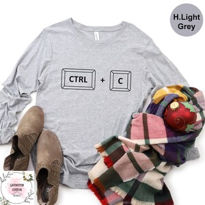 May include: Light grey long-sleeve shirt with the text "CTRL + C" in black. The shirt is paired with brown suede boots and a colorful plaid scarf. A red and gold Christmas ornament is also visible.