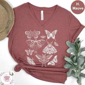 a red v-neck t-shirt with a white graphic design of various butterflies printed on it. The t-shirt is placed on a white surface, and there is a hat and some greenery visible in the background.