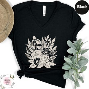 a black v-neck t-shirt with a white floral design, placed on a white surface next to a hat and a plant.