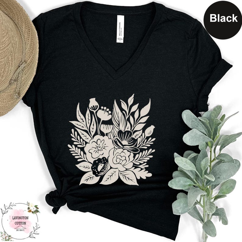 a black v-neck t-shirt with a white floral design, placed on a white surface next to a hat and a plant.