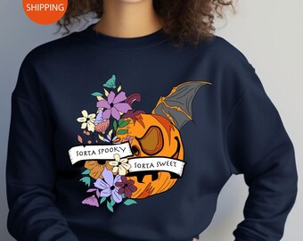 May include: A navy blue crewneck sweatshirt featuring a graphic of a split pumpkin with one bat wing and a cluster of colorful flowers. A white banner across the design displays the text "SORTA SPOOKY" on the left and "SORTA SWEET" on the right.