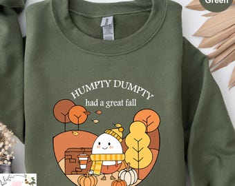 May include: Military green sweatshirt with a fall-themed graphic. The design features an egg character with a scarf, hat, and coffee cup, surrounded by pumpkins and trees. The text "HUMPTY DUMPTY had a great fall" is printed above the graphic.