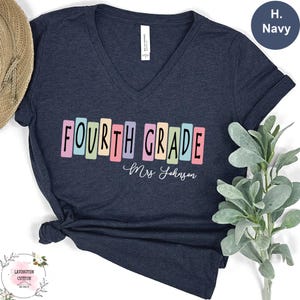 May include: Navy blue v-neck t-shirt with the words "FOURTH GRADE" in colorful block letters. Below the text is the name "Mrs. Johnson" in cursive. The shirt is made of soft material.