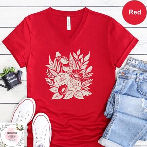 a red V-neck t-shirt with a white floral design, a pair of blue jeans, white sneakers, and a black mug, all arranged on a white wooden surface.