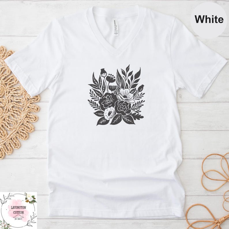 A white t-shirt with a black floral graphic printed on it, placed on a white wooden surface.