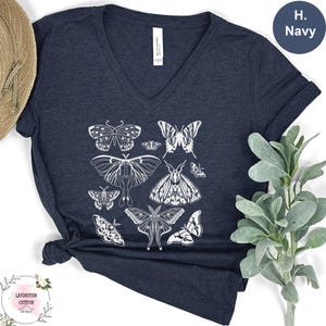 a navy blue t-shirt with a graphic design of various butterflies printed on it. The t-shirt is placed on a white surface, and there is a plant or greenery to the right of the t-shirt.