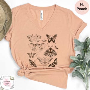 a peach-colored t-shirt with a graphic design of various butterflies printed on it. The t-shirt is placed on a white surface, and there is a plant visible in the background.