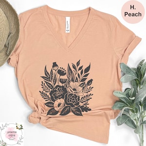 a peach-colored t-shirt with a black floral design, placed on a white background. There is a straw hat and some greenery, such as a plant, to the side of the t-shirt.