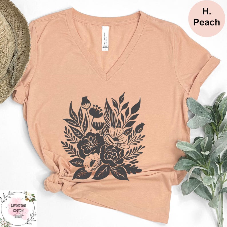 a peach-colored t-shirt with a black floral design, placed on a white background. There is a straw hat and some greenery, such as a plant, to the side of the t-shirt.
